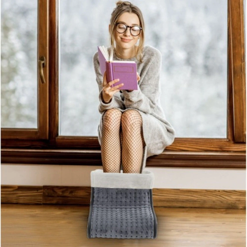 Mladá žena sedí s knihou u okna s nohama v elektrickém ohřívači na nohy.